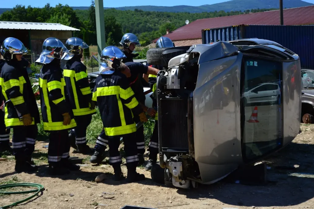 Firefighters in reflective helmets and yellow-striped uniforms surround a silver car flipped on its side, suggesting an accident scene on a sunny day.