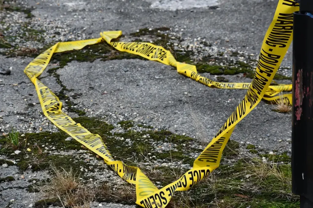 Yellow police tape forms a heart shape on cracked pavement, surrounded by moss and sparse grass, conveying a somber and mysterious tone.