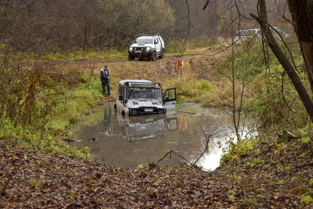 Off-road vehicle stuck in a muddy pond in a forested area, with the driver's door open. A person watches while another vehicle waits nearby.