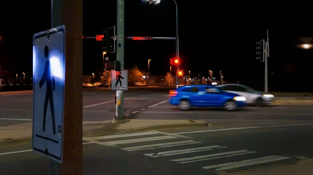Nighttime street scene with a visible pedestrian crossing sign in the foreground. A motion-blurred blue car speeds through an intersection under red traffic lights.