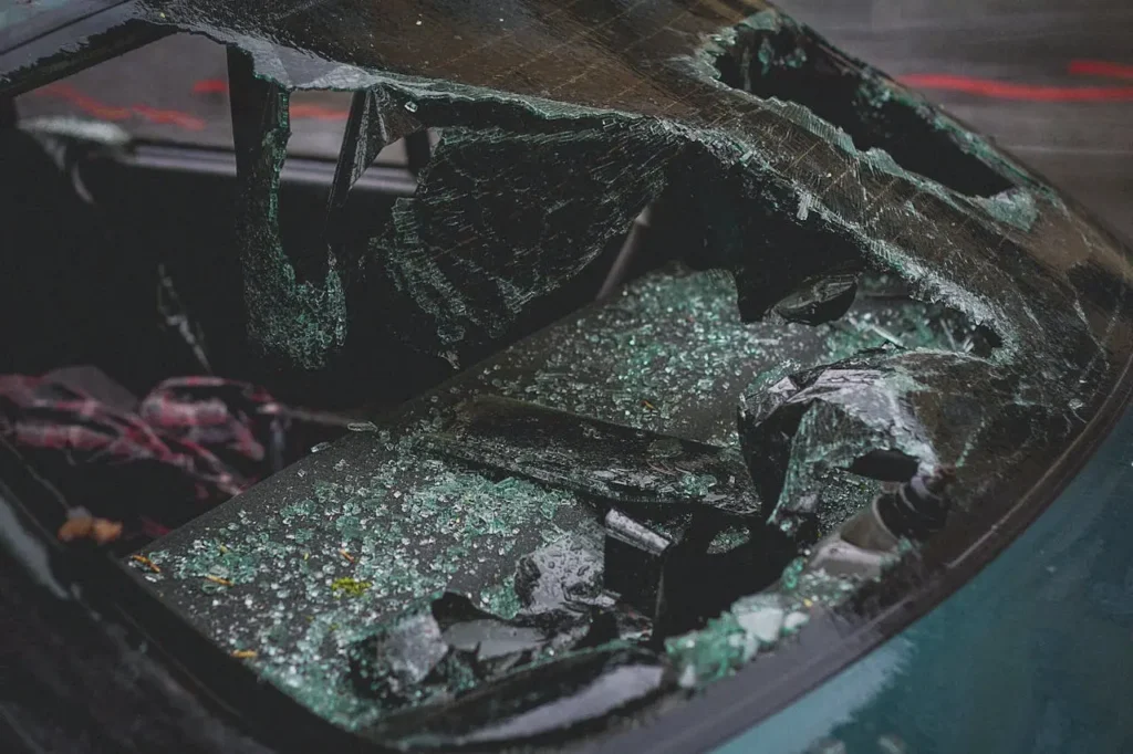 A close-up of a car windshield shattered into large and small pieces, with broken glass scattered inside the vehicle.