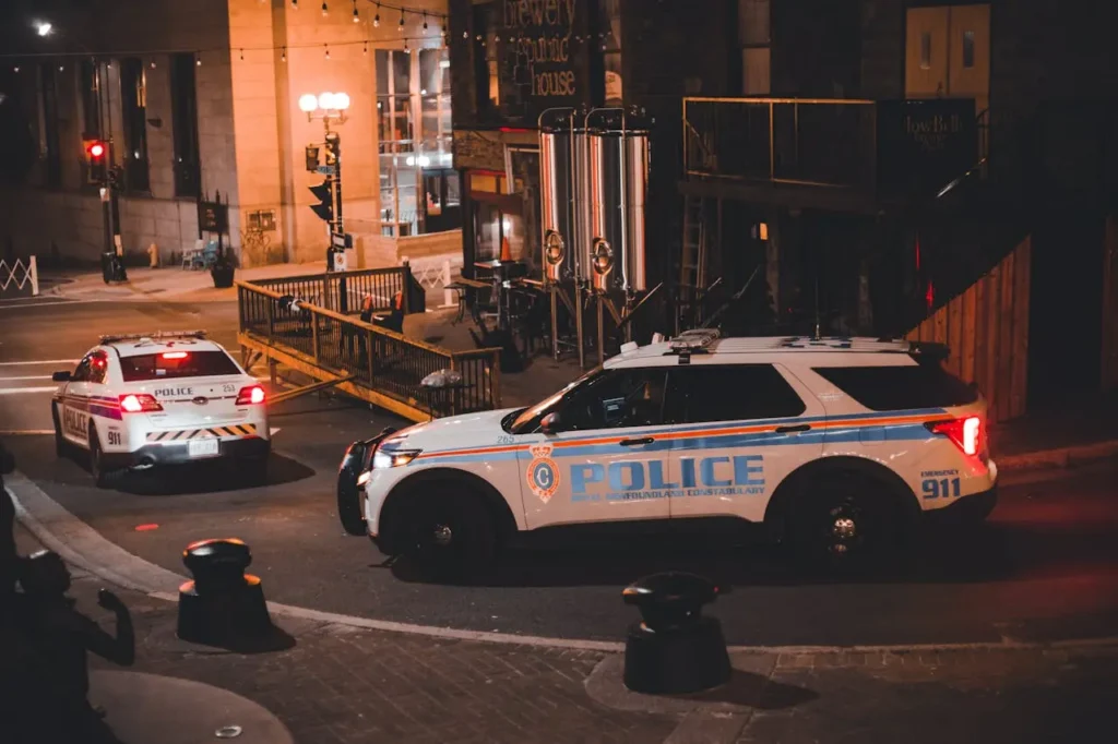 Two police SUVs with flashing lights are parked on a city street at night near a building with outdoor seating and string lights.