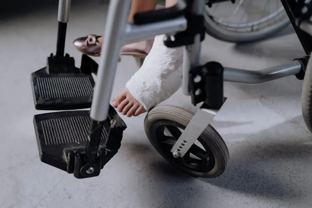 A close-up of a person in a wheelchair. One foot is bandaged, resting on a footrest. The other foot is in a pink shoe, showing a contrast of injury and normalcy.