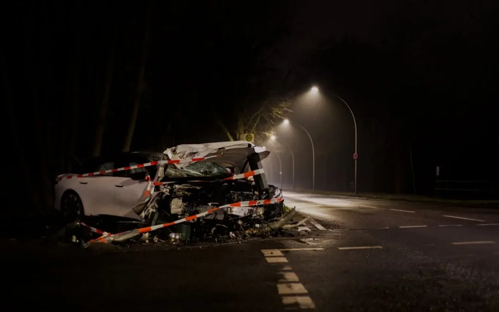 Severely damaged white car after a nighttime crash on a dark roadway, surrounded by police tape and illuminated by streetlights.