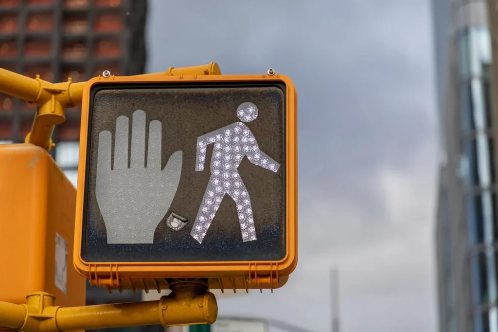 Traffic signal showing a white pedestrian figure next to an illuminated orange hand for stop. The background includes a cloudy sky and tall buildings.